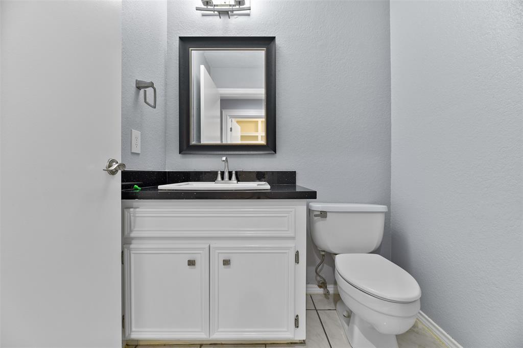 A half bathroom is conveniently located near the garage entrance of the home. Here, you'll find a black granite countertop with a rectangular basin sink white cabinetry, brushed nickel hardware, a framed mirror, stylish lighting, and tile floors.