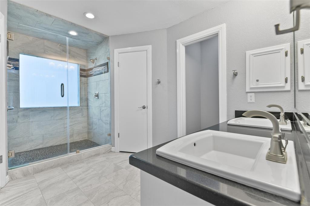 The en suite master bathroom has been masterfully renovated to include a sleek granite top vanity with dual rectangular basin sinks, white cabinetry, designer lighting, tile floors, his & hers closets, and an extra-large walk-in shower with a seamless glass door and a stylish tile surround.