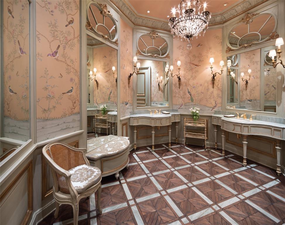The Ladies Powder Room features a lovely octagonal salon with charming wallpaper design over paneled wainscoting, two built in consoles with under mounted hand painted basin with reed and ribbon custom fixtures set into wall mounted marble top tapered wood legs and gilt accents and a pier mirror with an upholstered settee.
