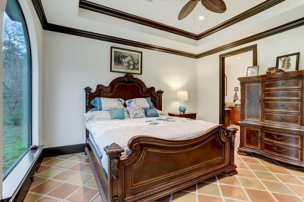 Indulge in comfort within this masterfully crafted bedroom, featuring a tray ceiling with a classic ceiling fan, large windows that frame the lush outdoors, and exquisite tile flooring. The room is a retreat, offering a serene view and a spacious environment for relaxation and repose.